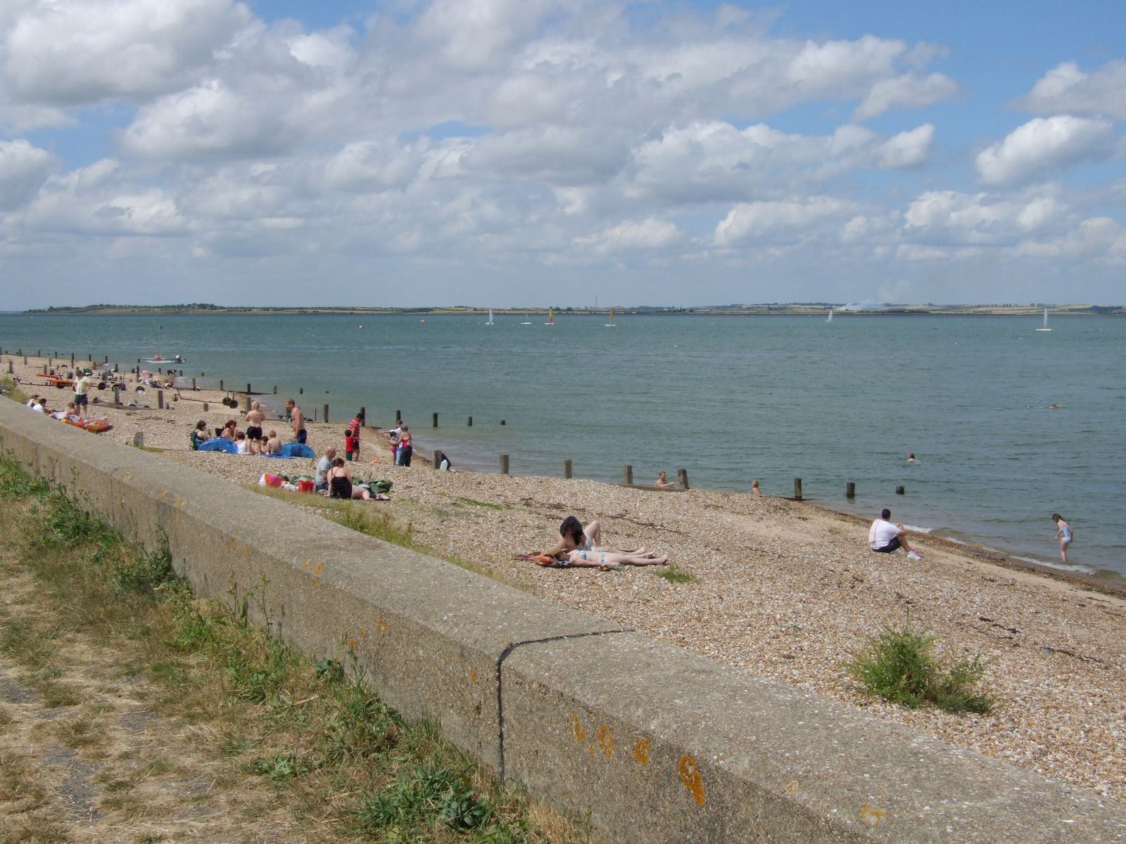 Families on the shingle at Seasalter beach on a sunny day, with the sea wall in the foreground