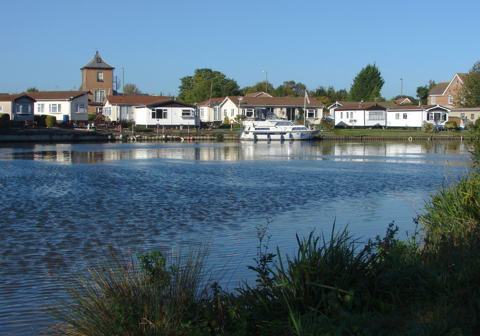 Single-storey park homes with lawns along the River Thames at Thames Retreat, with a motor cruiser moored at the bank